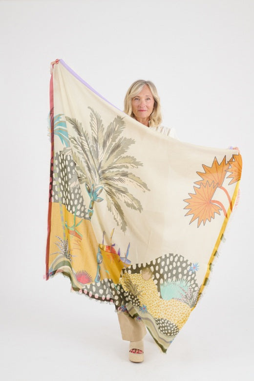 A woman with shoulder-length blonde hair models the STORIATIPIC MALI MAX Scarf in beige—a breathable cotton modal accessory with green, orange, and blue accents—paired with a cream embroidered blouse against a light gray background.
