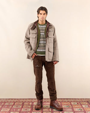 A man wearing the AMUNDSEN SPORTS - HUNTSMAN JACKET over a cream turtleneck sweater and tan pants stands facing the camera against a white background.