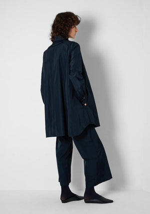 A person with curly hair wears the KATHARINA HOVMAN oversized taffeta blouse, featuring mother-of-pearl buttons, along with black pants. They stand in a minimalist setting with hands in pockets, gazing at the camera.