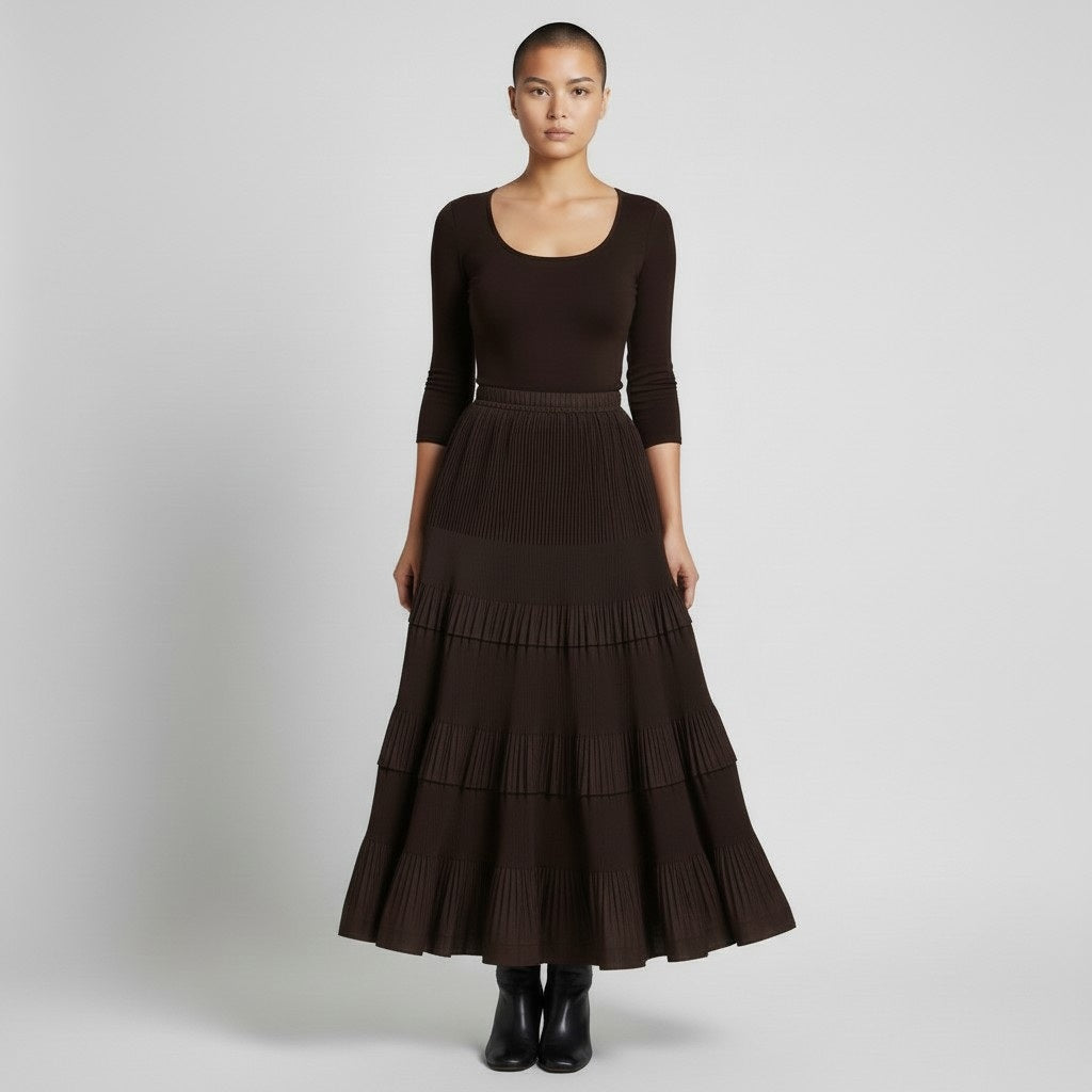 A woman with a shaved head and neutral expression wears the MEIMEIJ RUFFLE SKIRT IN MOCHA, styled with a fitted long-sleeve brown top and black ankle boots, standing against a plain background.