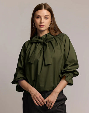 A woman with long brown hair models the HINSON WU ALINA LONG SLEEVE SHIRT, a bright blue blouse with a bow neck and ruffled sleeves, paired with dark pants. She stands against a light background, looking at the camera.