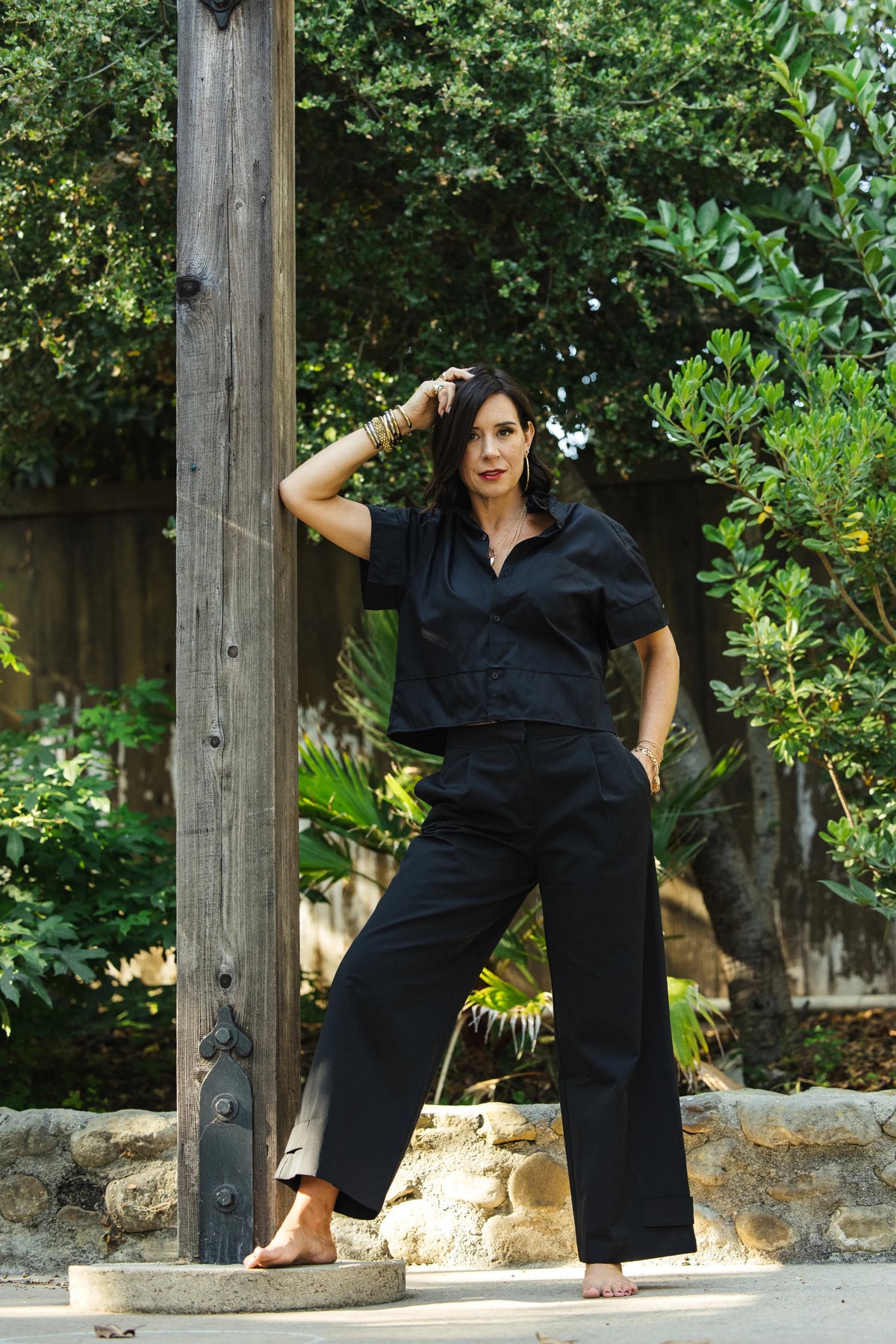 A woman in a black KMJ REESE SHIRT stands barefoot outdoors, leaning against a wooden post with one hand on her hip. She poses confidently, surrounded by lush green plants and trees.