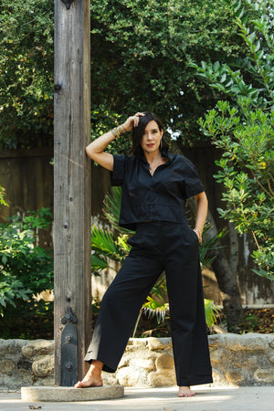A woman in a black KMJ REESE SHIRT stands barefoot outdoors, leaning against a wooden post with one hand on her hip. She poses confidently, surrounded by lush green plants and trees.