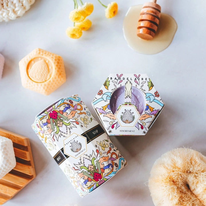 Four pastel-colored, triple-milled soaps—lavender, pink, white, and orange—are stacked next to a decorative floral-patterned box labeled “BAUDELAIRE - HONEY SOAP ASSORTMENT 1.4 OZ QUARTET” by BAUDELAIRE.