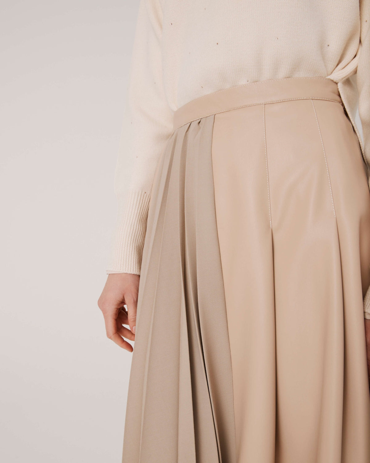 A person with long hair wearing sunglasses poses against a plain background. They wear a cream-colored sweater, a SFIZIO pleated mix media skirt in beige faux leather, and matching knee-high snakeskin boots. They hold a snakeskin-patterned handbag.