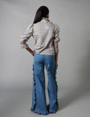 A woman models the HOPE FOR FLOWERS - FRILLED BUTTON DOWN SHIRT by HOPE FOR FLOWERS, made from GOTS certified organic cotton, with a high collar and wide sleeves, paired with flared blue jeans featuring fringe along the side seams.