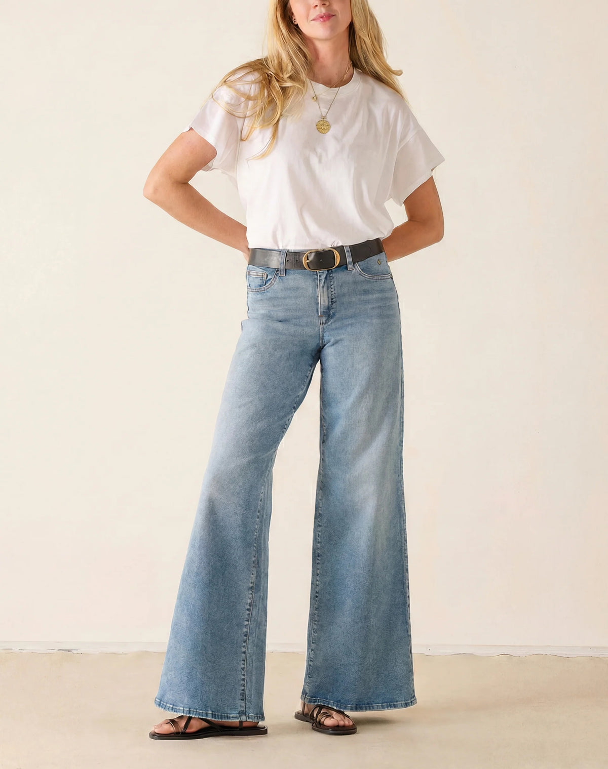 A woman stands against a plain light background wearing a white t-shirt, black sandals, and the MERSEA KATE Wide Leg Jeans in Cloud Nine—a relaxed fit, light blue four-way stretch denim style. Her head is turned away as she steps forward with one leg.
