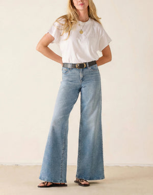 A woman stands against a plain light background wearing a white t-shirt, black sandals, and the MERSEA KATE Wide Leg Jeans in Cloud Nine—a relaxed fit, light blue four-way stretch denim style. Her head is turned away as she steps forward with one leg.