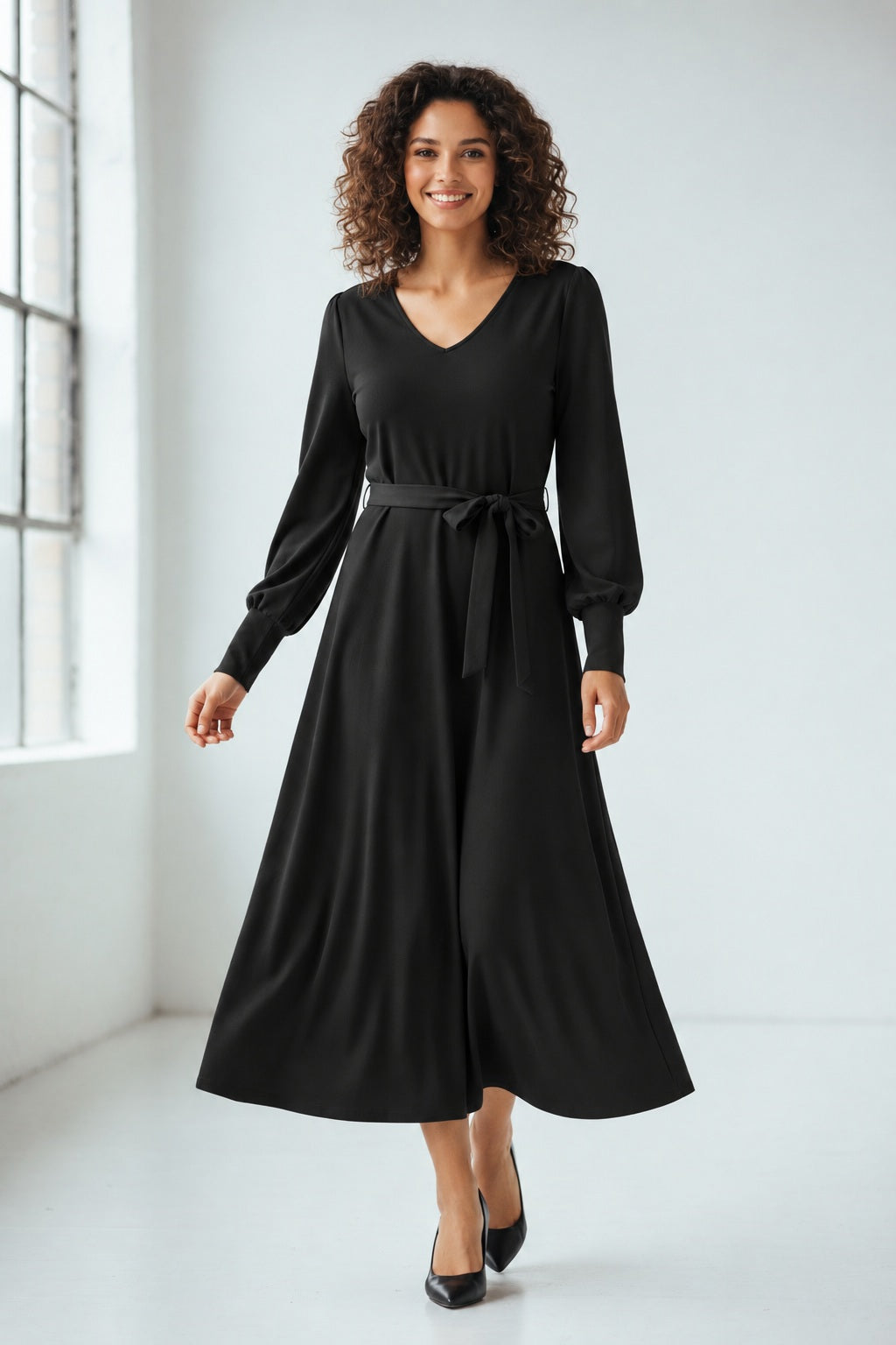 A woman with long, light brown hair stands against a plain background. She is wearing the SFIZIO long sleeve v-neck flare skirt dress, featuring a tie waist. Looking to her left with a calm expression, she has hoop earrings and exudes minimalist style.