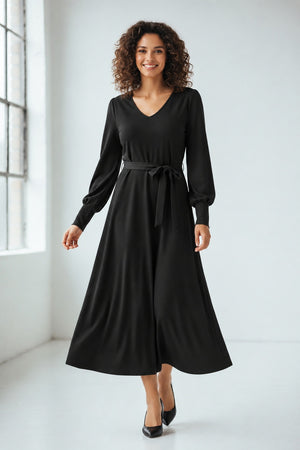 A woman with long, light brown hair stands against a plain background. She is wearing the SFIZIO long sleeve v-neck flare skirt dress, featuring a tie waist. Looking to her left with a calm expression, she has hoop earrings and exudes minimalist style.