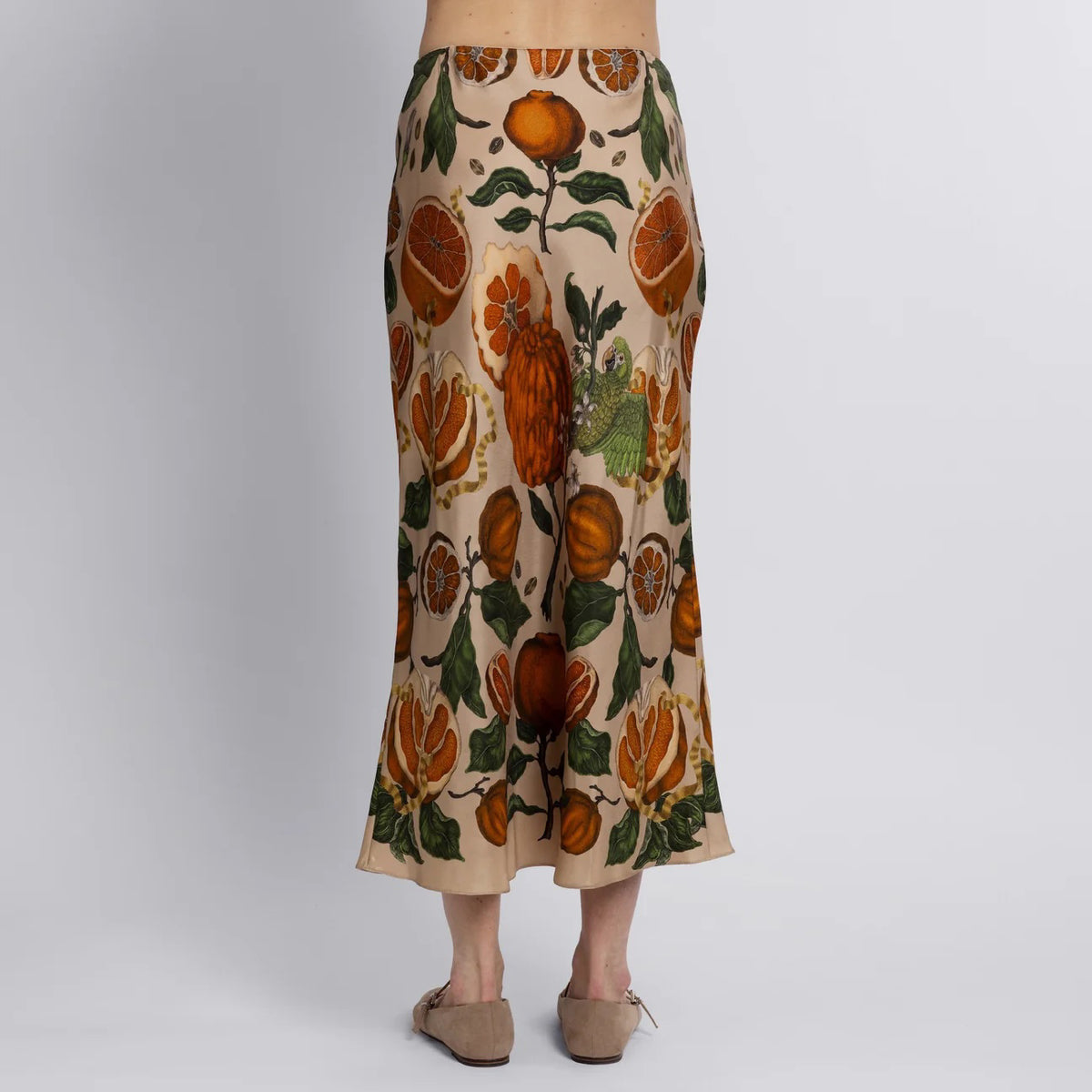 A person stands barefoot on a wooden floor wearing the SABINA SAVAGE - ORANGES AND LEMONS SILK BIAS SKIRT from SABINA SAVAGE ENGLAND, featuring an orange and green floral and fruit exotic print. The plain white background highlights the skirt&#39;s elegant bias cut.