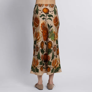 A person stands barefoot on a wooden floor wearing the SABINA SAVAGE - ORANGES AND LEMONS SILK BIAS SKIRT from SABINA SAVAGE ENGLAND, featuring an orange and green floral and fruit exotic print. The plain white background highlights the skirt's elegant bias cut.