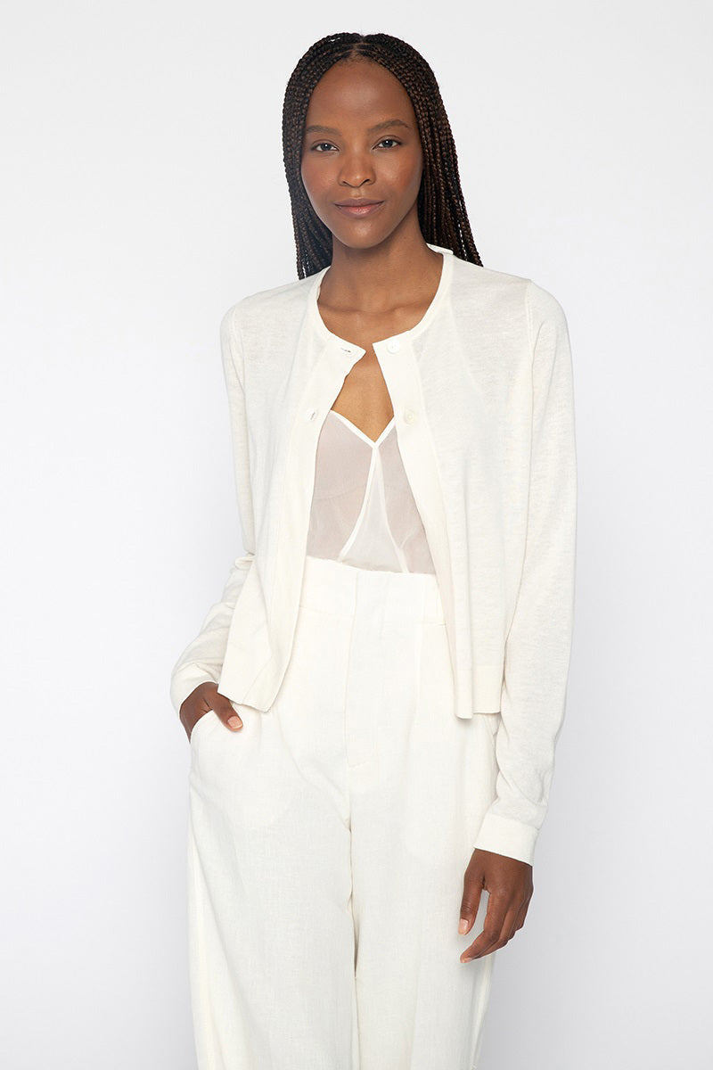 A woman with long braided hair stands against a plain background, wearing an all-white outfit. She has on the KINROSS CASHMERE - DRESS CARDIGAN by KINROSS, layered over a semi-sheer silk top and high-waisted trousers. Her left hand is in her pocket, and she has a gentle smile.