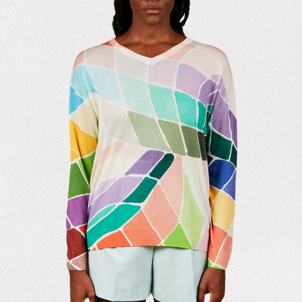 A person wearing the IN BED WITH YOU - BATWING SLEEVE V NECK JUMPER stands against a plain, light-colored background. The colorful geometric pattern featuring squares and circles in shades of pink, purple, red, yellow, and white adds flair to this stylish piece by the Italian brand IN BED WITH YOU/CLAUDIOLAI SRL. The person is visible from the shoulders down to the waist.
