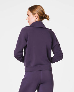 A person with short hair is wearing a SPANX AIRESSENTIALS HALF ZIP sweater in navy blue, made from lightweight spacer fabric, paired with black pants. Their arms are crossed over their chest as they stand against a plain, light grey background.