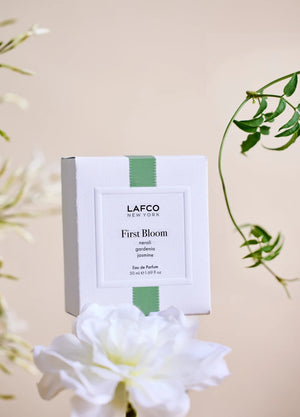 A box of LAFCO - FINE FRAGRANCE, FIRST BLOOM 50ML stands among white flowers and green vines against a pale beige background.