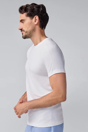 A man with short dark hair and a beard wears a slim fit white MACK WELDON - 18 HOUR JERSEY CREWNECK UNDERSHIRT and light blue pants, standing in profile against a plain light gray background.