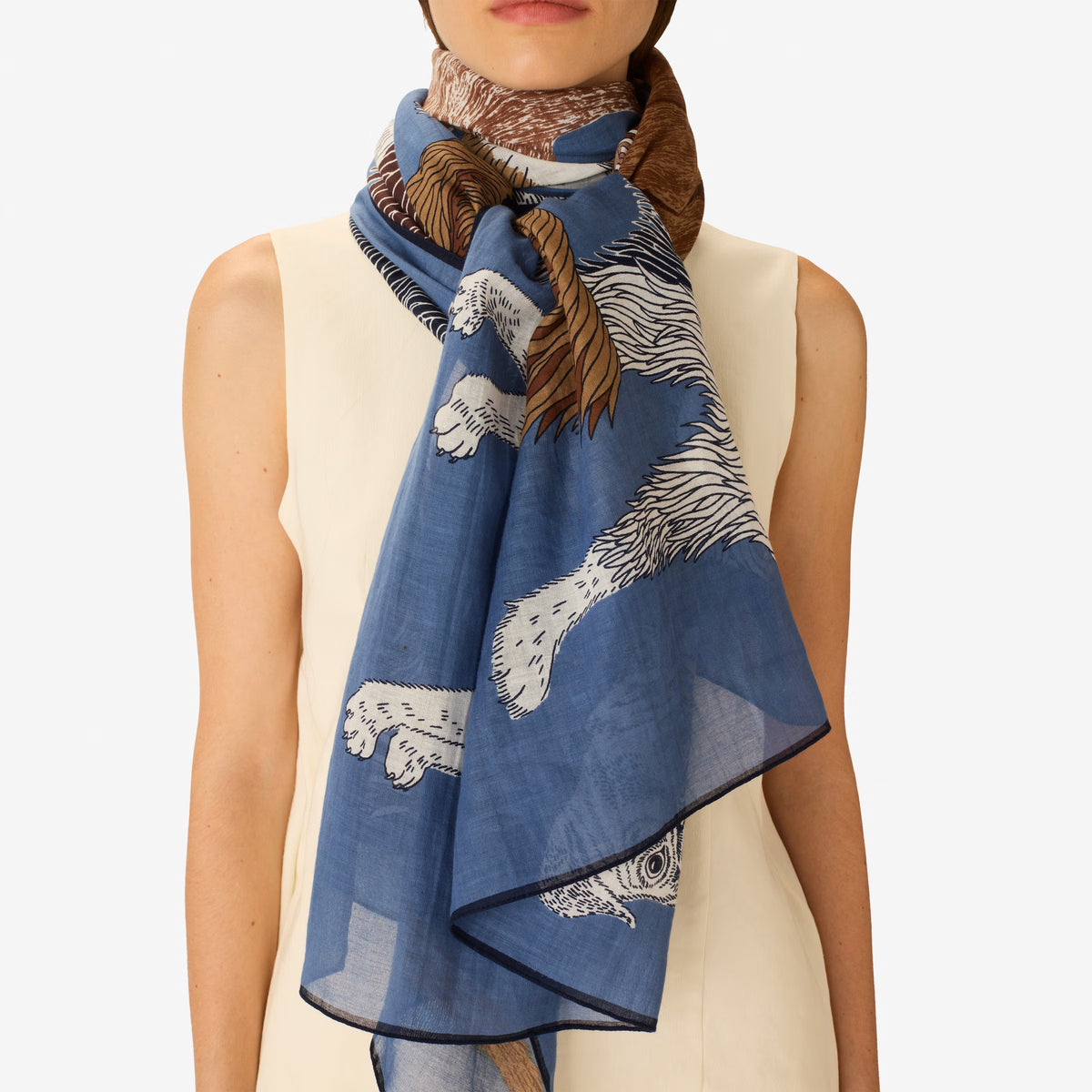 A person wears a sleeveless beige top and the INOUI EDITIONS - TOUTOU SCARF IN BLUE by INOUI, featuring a detailed animal print with white fur and brown accents, wrapped around their neck against a plain white background.