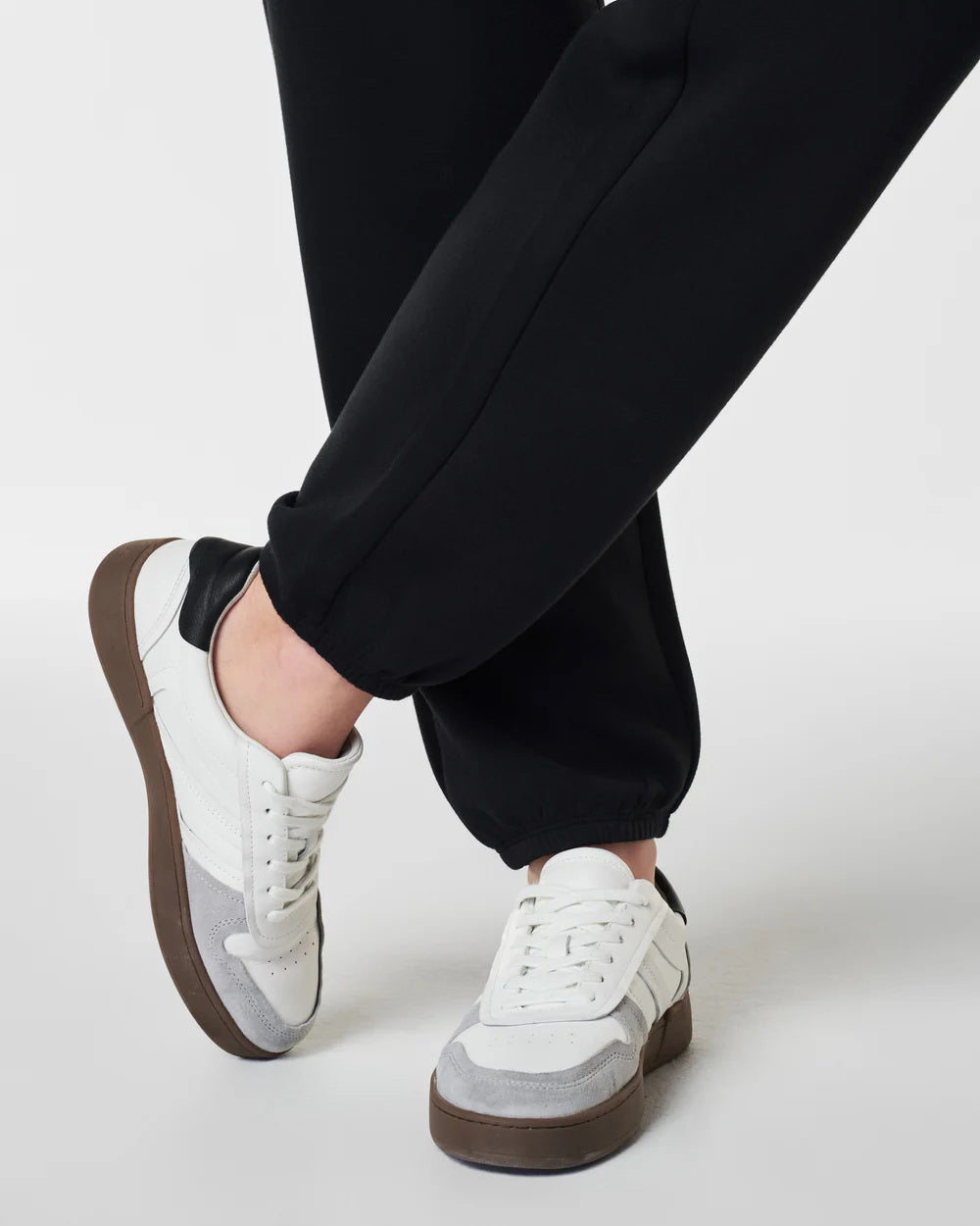 Dressed in SPANX - ARESSENTIALS JOGGER pants crafted from lightweight fabric, a person stands with hands in pockets, pairing them with a matching hoodie. They complete their look with white sneakers featuring brown soles. The plain white background accentuates their outfit&#39;s style and comfort.