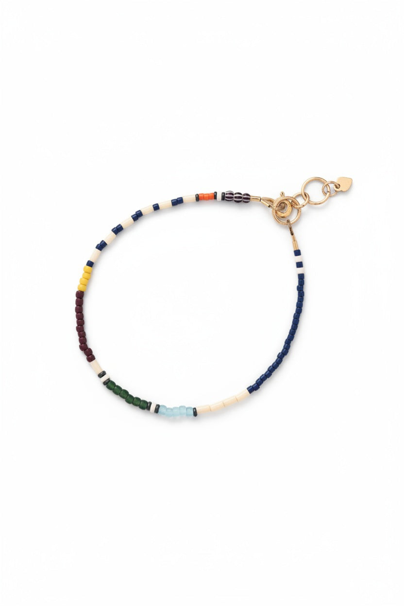 The ALICE RISE Thinnest Line Bracelet in Navy and Olive, featuring Japanese glass beads and gold clasps, is displayed on a blue textured surface with a large seashell partially visible.
