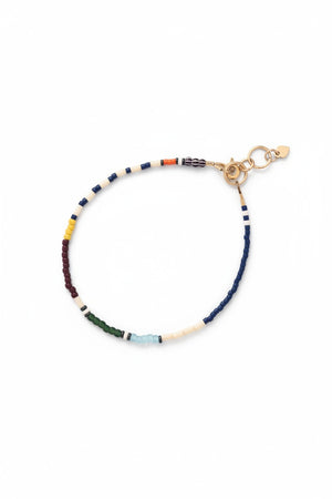 The ALICE RISE Thinnest Line Bracelet in Navy and Olive, featuring Japanese glass beads and gold clasps, is displayed on a blue textured surface with a large seashell partially visible.