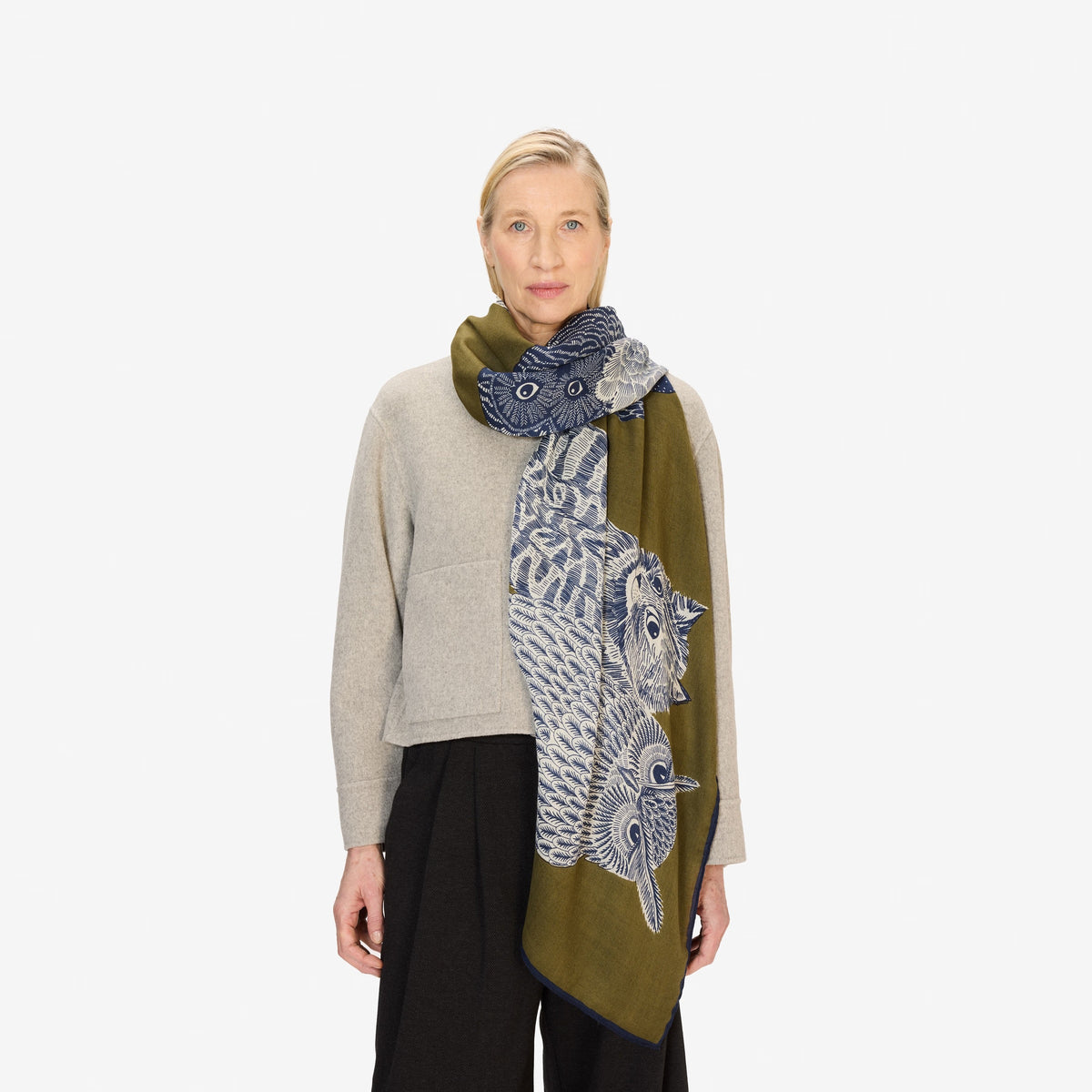 A woman stands against a white background, wearing a beige long-sleeve top, black wide-leg pants, and the INOUI EDITIONS - Owls on a Branch Wool Scarf in Khaki by INOUI, draped elegantly over her shoulder.
