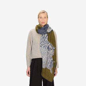 A woman stands against a white background, wearing a beige long-sleeve top, black wide-leg pants, and the INOUI EDITIONS - Owls on a Branch Wool Scarf in Khaki by INOUI, draped elegantly over her shoulder.