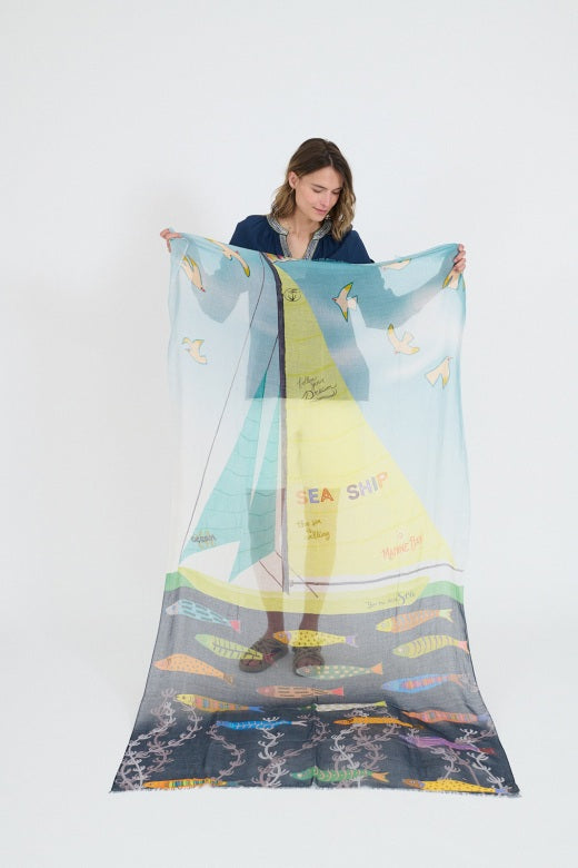 A woman with shoulder-length wavy hair models the STORIATIPIC - GERTRUD SCARF, styled around her neck over a navy blue top, against a plain white background.