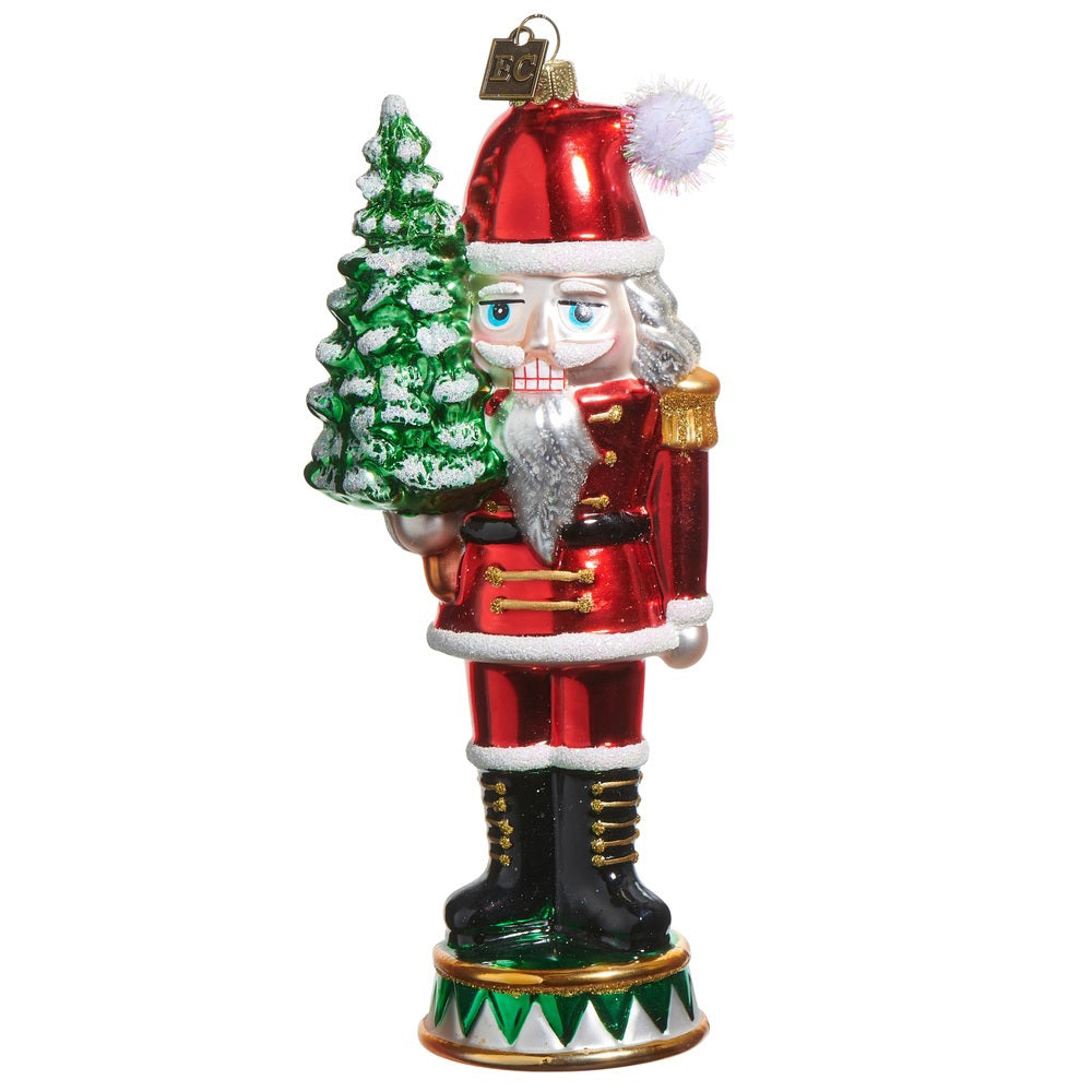 The RAZ IMPORTS NUTCRACKER SANTA ORNAMENT, in a red and gold uniform with a Santa hat, holds a snow-covered tree and stands on a round base beside a charming glass vase.