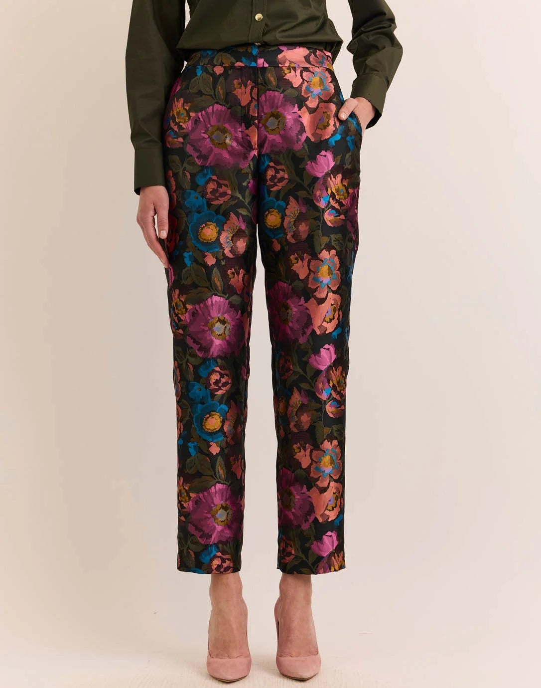 A person wears the HINSON WU Jolene Jacquard Pant in Floral Bouquet—high-waisted, ankle-length tailored trousers featuring a vibrant floral pattern in pink, purple, blue, and orange—paired with a dark green shirt and light pink heels.