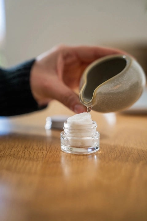 A hand pours liquid from a small ceramic pitcher into a glass jar filled with a white, fluffy substance atop a wooden surface, demonstrating the preparation of essential oil tablets for use with the MADISON AND GREEN - REST YOUR EYES AT THE DESK aromatherapy eye covers or reusable eye mask by MADISON GREEN.