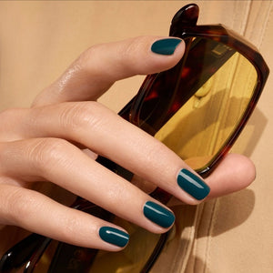 A hand with dark green painted nails rests on silver fabric, next to a matching bottle of MANUCURIST - GREEN™ Nail Polish in Dark Clover by MANUCURIST INC.