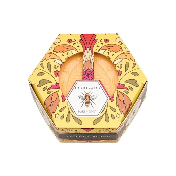 A luxurious BAUDELAIRE Pure Honey Soap 3.5 oz, featuring a hexagonal honeycomb pattern and beehive design, rests beside its floral BAUDELAIRE packaging. Nearby, a honey dipper releases golden honey over a textured backdrop. This exquisite soap is proudly made in the USA.