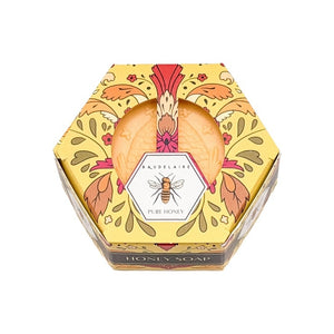 A luxurious BAUDELAIRE Pure Honey Soap 3.5 oz, featuring a hexagonal honeycomb pattern and beehive design, rests beside its floral BAUDELAIRE packaging. Nearby, a honey dipper releases golden honey over a textured backdrop. This exquisite soap is proudly made in the USA.