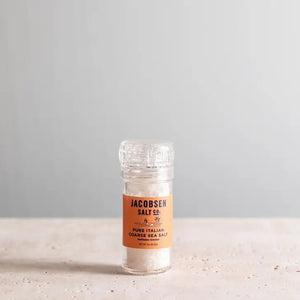 A clear plastic grinder of JACOBSEN SALT CO. GRINDER COARSE ITALIAN SEA SALT sits on a light surface with a plain gray background. The grinder features an orange label with black text.