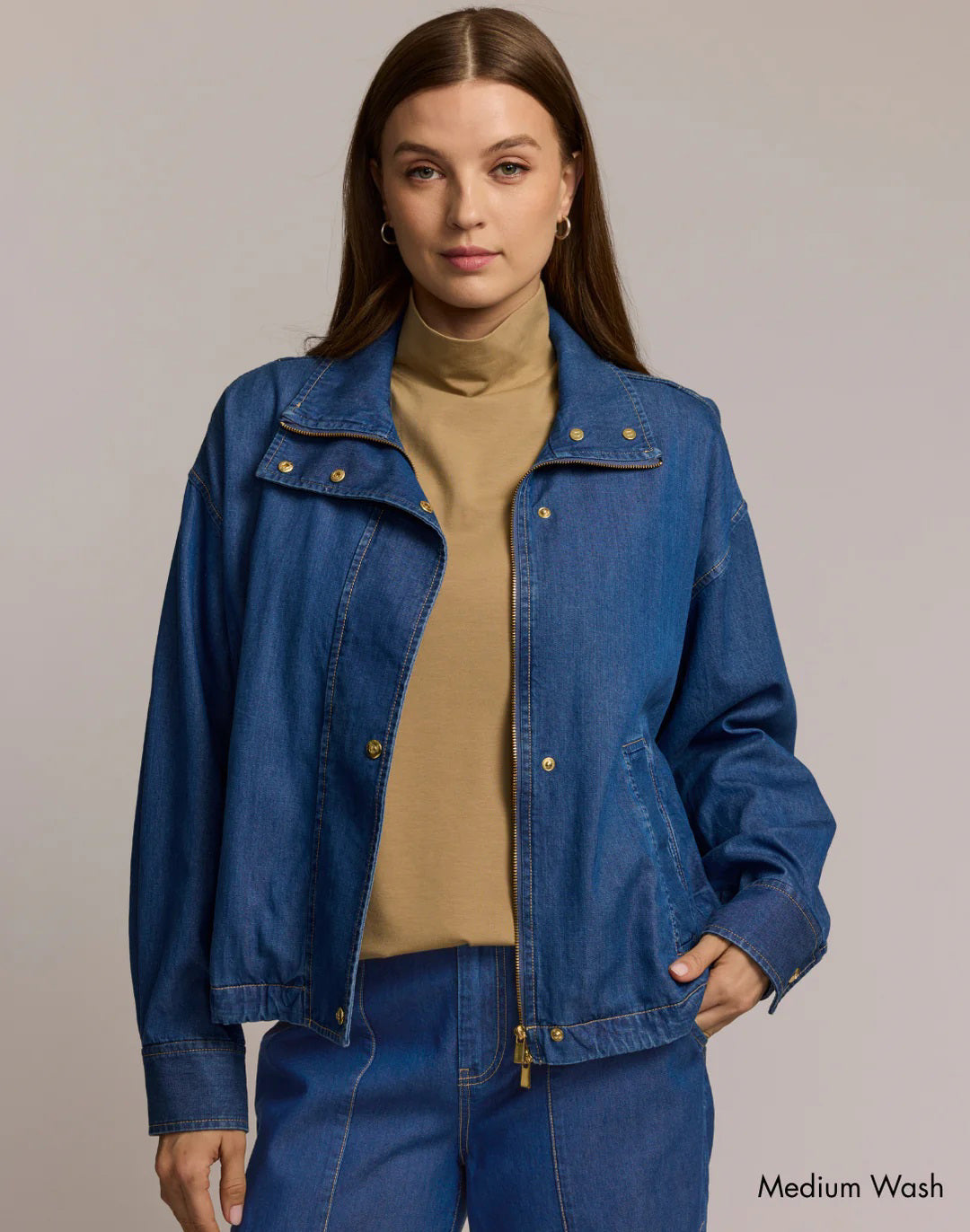 A woman wears the HINSON WU - LOUISE DENIM JACKET IN MEDIUM WASH over a tan turtleneck and blue denim pants, posing against a beige background. “Medium Wash” appears in the lower right corner.