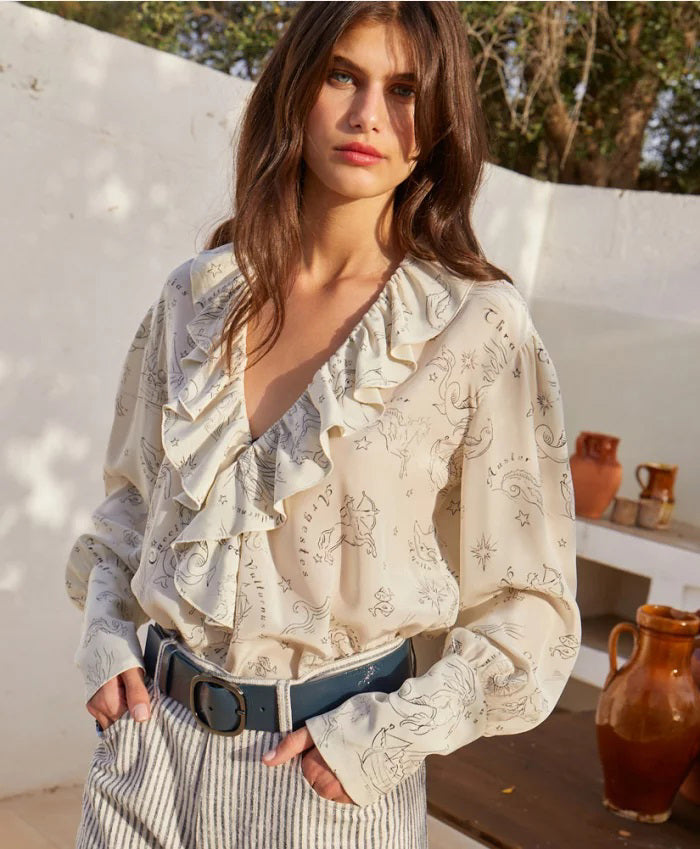 A woman with long brown hair stands outdoors, wearing the MOMONI RUFFLED BLOUSE and striped pants. She looks over her shoulder, with clay jugs and a wooden table in the background against a white wall.