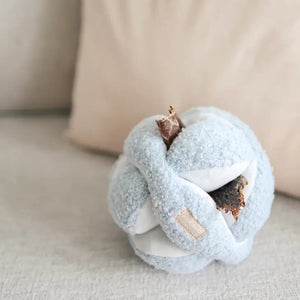 A small, light-colored dog stands on a light gray sofa, engaged in nosework play with the LAMBWOLF COLLECTIVE Monti Dog Toy in Oat. The background is simple and minimalistic.