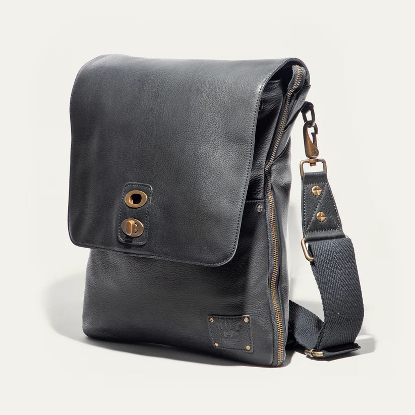 The WILL LEATHER GOODS OTTO Crossbody Bag in black full-grain leather features a front flap, gold-tone hardware, adjustable shoulder strap, and a small stitched patch on the lower right corner. Displayed against a plain white background.