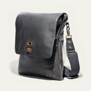 The WILL LEATHER GOODS OTTO Crossbody Bag in black full-grain leather features a front flap, gold-tone hardware, adjustable shoulder strap, and a small stitched patch on the lower right corner. Displayed against a plain white background.