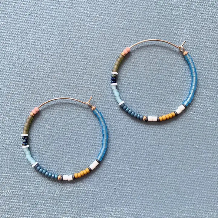 The ALICE RISE - BIG COLORLOOP EARRINGS IN SPRINGWATER by ALICE RISE feature two large hoops adorned with small, colorful Japanese glass beads, elegantly arranged and displayed on a textured light grey surface.