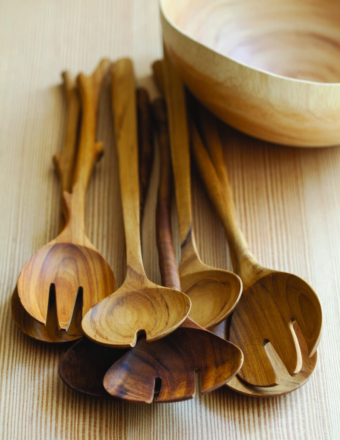 The BEHOME - TEAK SERVING SET by BE HOME, INC features two handcrafted teak servers—a spoon and a three-pronged fork—arranged crossed on a light blue background.
