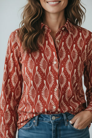 A woman with wavy brown hair smiles as she poses with one hand in her pocket, wearing the Hartford Cinema Shirt in Red Peacock Print—a loose-fitting, 100% cotton shirt featuring a vibrant red hue with a white peacock pattern—paired stylishly with blue jeans.