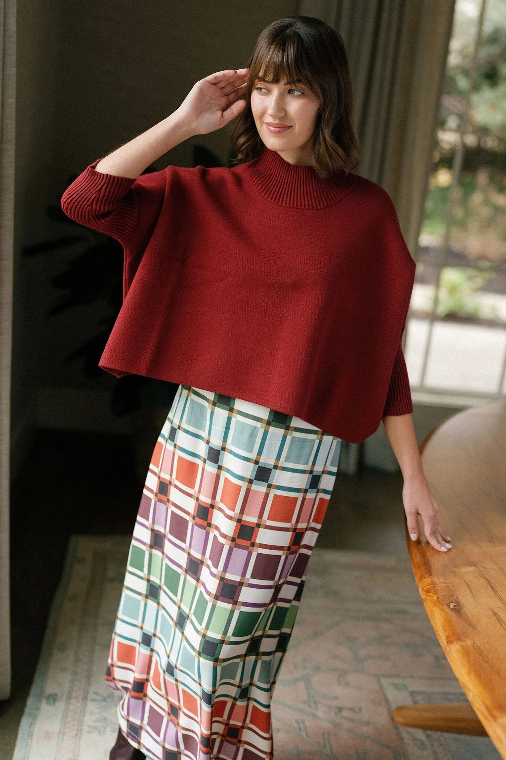 A woman stands indoors, wearing the BRITON COURT - SOPHIA OVERSIZED ONE SIZE SWEATER IN MULLBERRY by BRITON COURT, styled with a colorful plaid skirt. She smiles softly and touches her head with one hand, as natural light filters in from a window behind her.