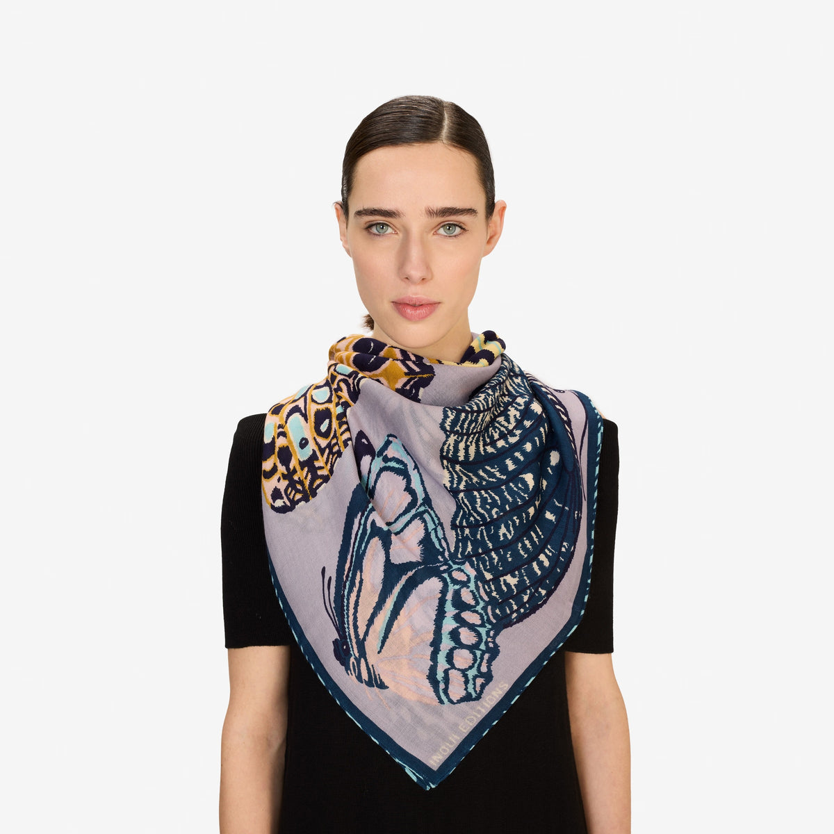 A woman with straight brown hair pulled back wears a dark top and drapes the INOUI EDITIONS PAPILLON (BUTTERFLIES) Square Scarf in Grey by INOUI, featuring abstract and butterfly patterns, around her neck against a white background.