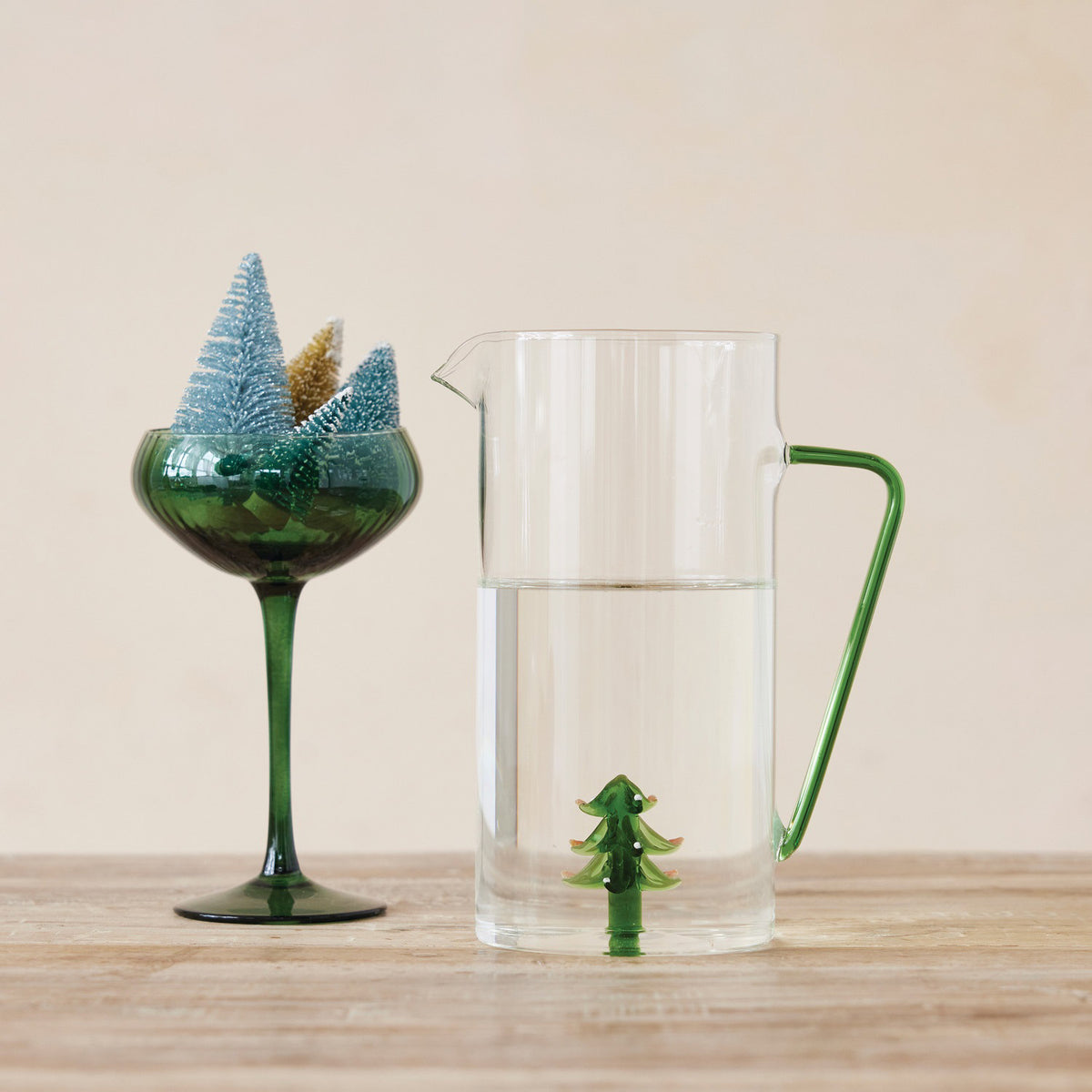 The CREATIVE COOP GLASS PITCHER WITH HOLIDAY TREE features a clear design, a green handle, and a small glass holiday tree inside the bottom.