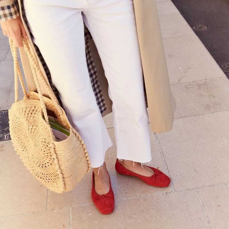 A person in white jeans, a beige coat, and carrying a round straw bag stands on light stone pavement, wearing REQINS HARMONY suede ballerina flats in rouge by REQINS.