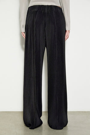 Side view of a person wearing the MEIMEIJ Long Pleated Pant in black, highlighting the elegant texture of pleated fabric. The pants are paired with a light gray, long-sleeved top, and the ensemble's draping is well highlighted against a plain background and light-colored floor.