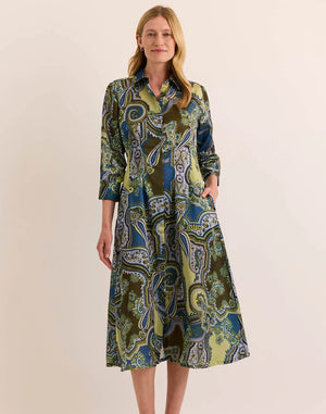 A woman stands smiling against a plain beige background, wearing the HINSON WU Martina 3/4 Sleeve Opulent Paisley Print Dress—a long-sleeve, collared, button-down dress featuring a green, blue, and brown paisley pattern and crafted from luxurious Italian polyester fabric for a fluid drape and silky hand feel.