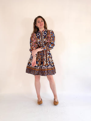 A woman poses against a plain background in the FARYLINA ANDI DRESS, a colorful bohemian style with long puff sleeves and a brown belt, styled with tan flats and long hair, one leg crossed in front of the other.